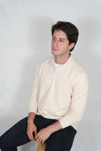 A man wearing a cream-colored long-sleeve polo shirt and dark pants, sitting against a plain light gray studio background.
