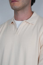 Close-up of a man wearing a light cream-colored long-sleeve polo shirt over a white undershirt, against a plain light gray background.