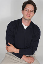 A man wearing a dark navy long-sleeve polo shirt over a white t-shirt and beige pants, sitting against a light gray background.