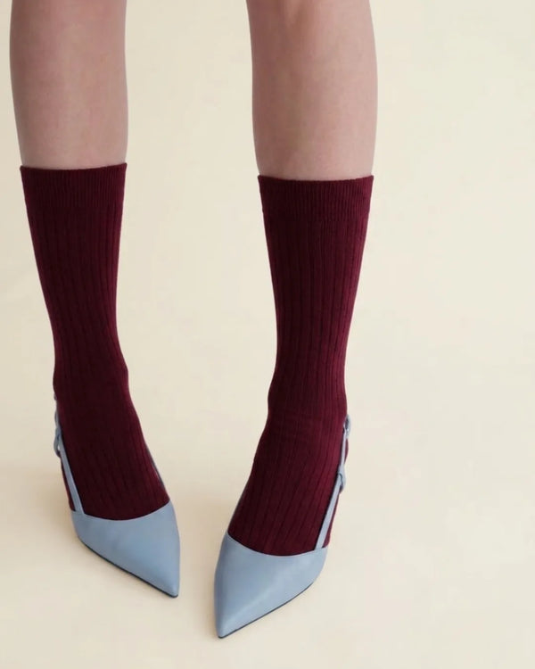 A person's legs wearing bordeaux ribbed crew socks with light blue pointed-toe heels, shown against a plain beige background.
