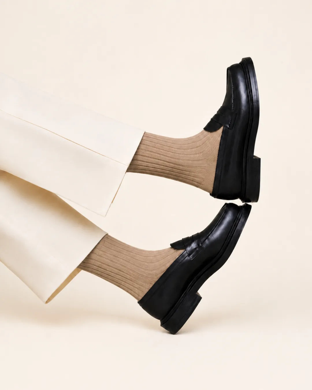 A person wearing dark leather penny loafers with latte-colored ribbed socks and cream trousers, shown from the knees down against a warm off-white studio background.