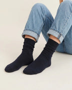 A person seated with legs crossed, wearing black ribbed crew socks and  cuffed jeans against a plain light studio background.