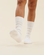 A close-up of a person's legs wearing cream ribbed crew socks, with one foot raised, against a solid light background.