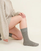 A person kneeling while wearing heather light grey crew socks with a knit sweater and shorts against a plain background.