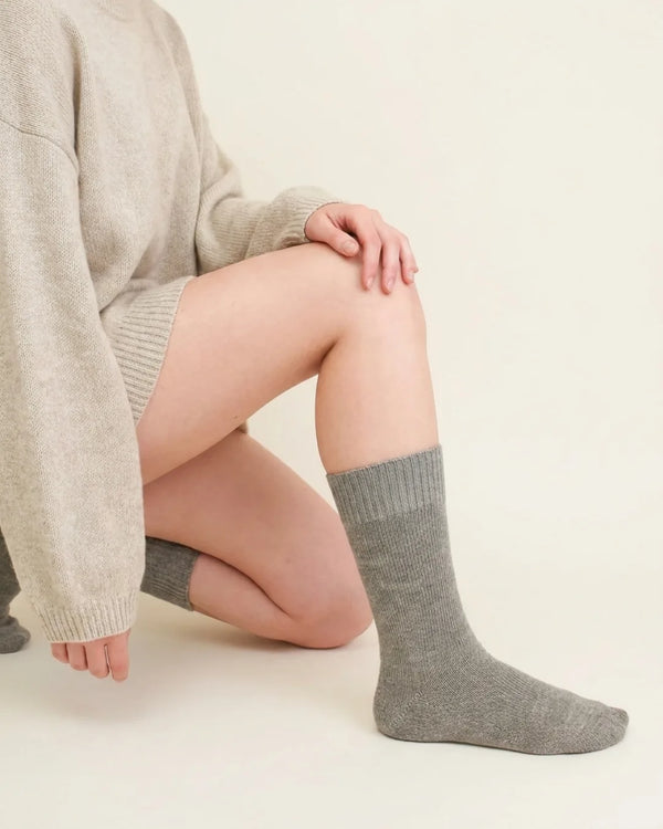 A person kneeling while wearing heather light grey crew socks with a knit sweater and shorts against a plain background.