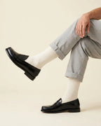A person seated with crossed legs wearing light gray cuffed trousers, panna ribbed socks, and dark leather penny loafers against a plain warm beige studio background.