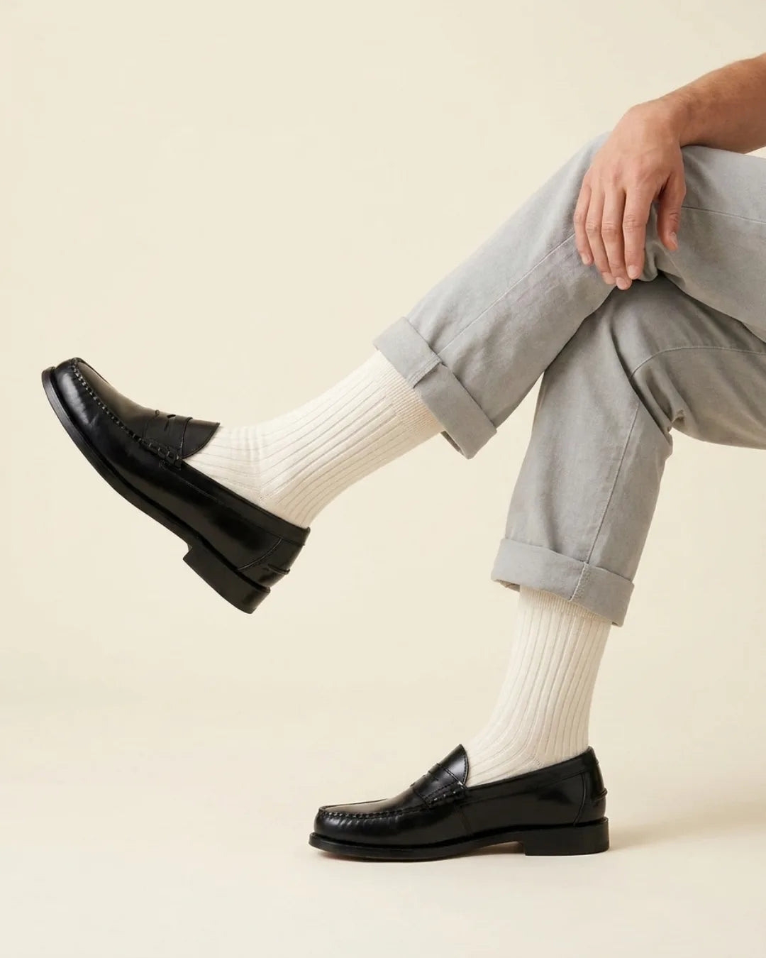 A person seated with crossed legs wearing light gray cuffed trousers, panna ribbed socks, and dark leather penny loafers against a plain warm beige studio background.