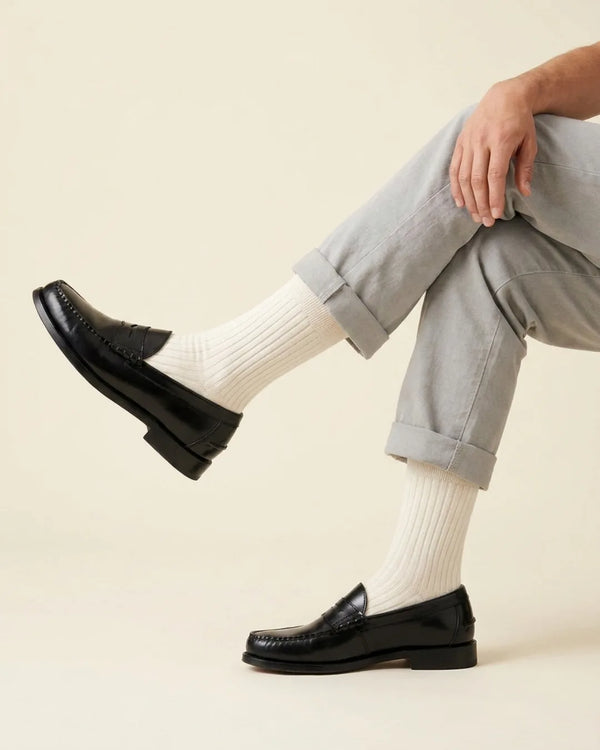 A person seated with crossed legs wearing light gray cuffed trousers, panna ribbed socks, and dark leather penny loafers against a plain warm beige studio background.