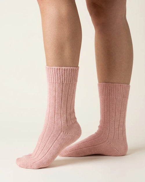 A pair of ribbed ankle socks in a dusty pink color, shown on a model's feet against a plain off-white background.